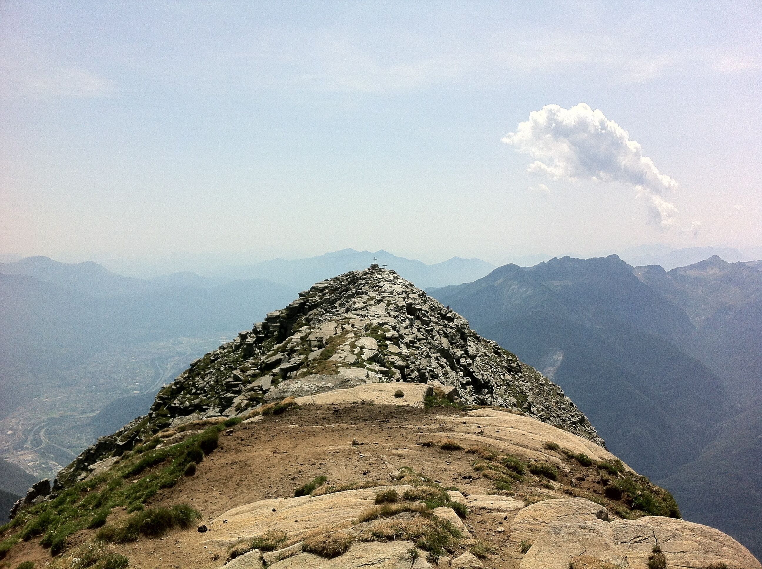 A rocky mountain peak with a cross at the summit, surrounded by a valley and distant mountains.