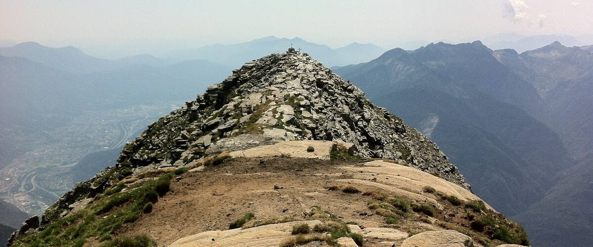 A rocky mountain peak with a cross at the summit, surrounded by a valley and distant mountains.