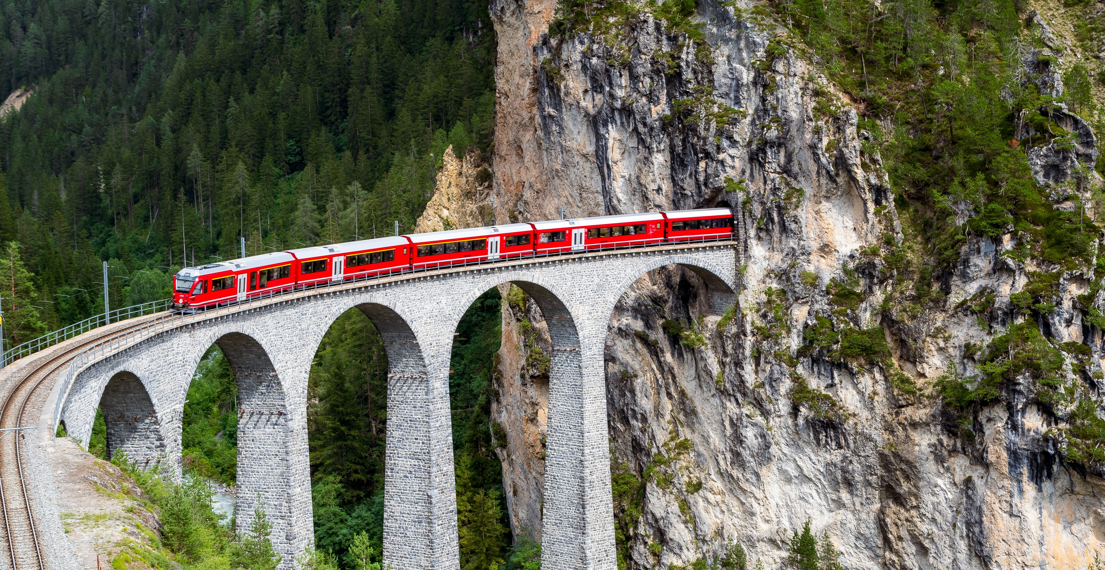 A red train on a stone bridge with multiple arches, surrounded by forested mountains.