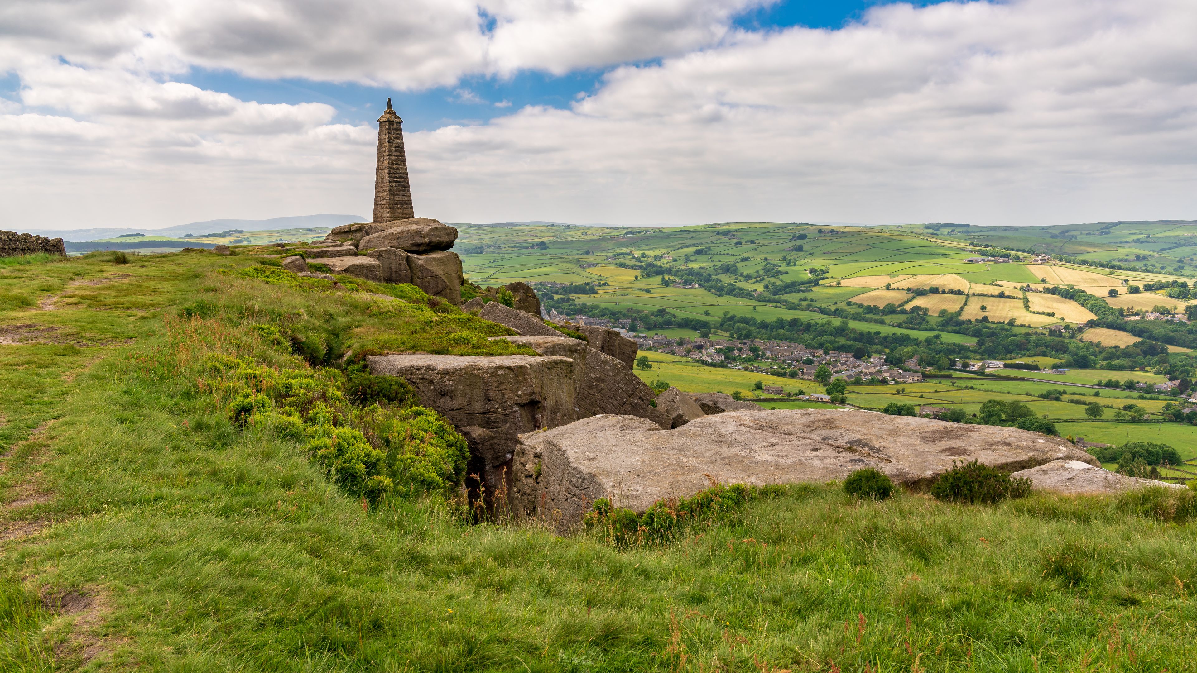 A stone monument on a grassy hill overlooking a valley with fields and a small village.