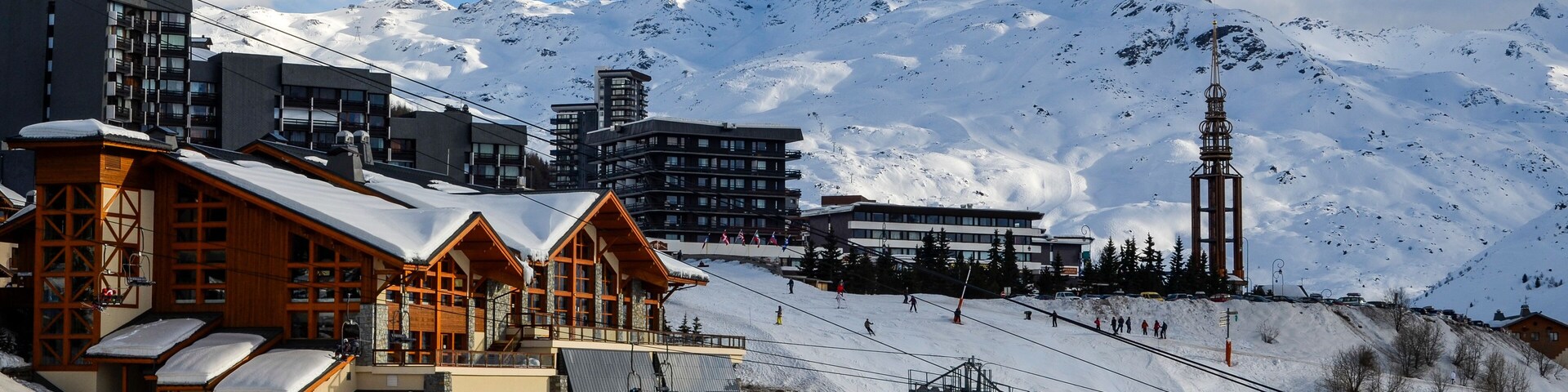 A ski resort with wooden buildings, a ski lift, and a snow-covered mountain in the background.