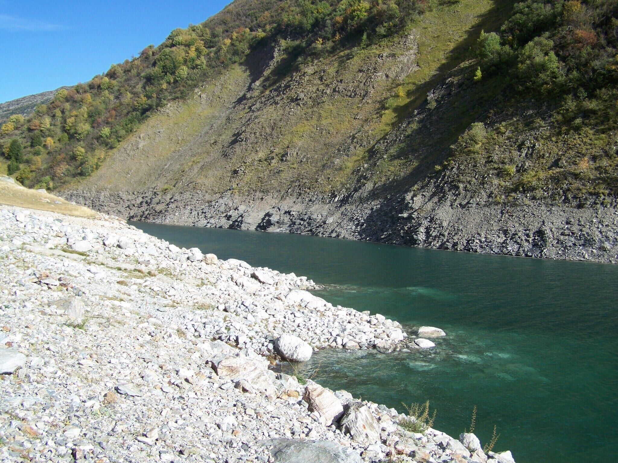 A rocky shoreline with clear water and a steep hillside in the background.