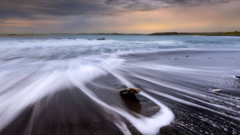 A beach with a smooth, flowing sand surface and a rock in the foreground.