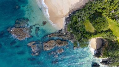 Aerial view of turquoise waters meeting the sandy shore near lush green headland, Langs Beach, Northland Region, New Zealand.