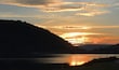 A sunset over a lake with a mountain in the foreground and a road leading towards it.