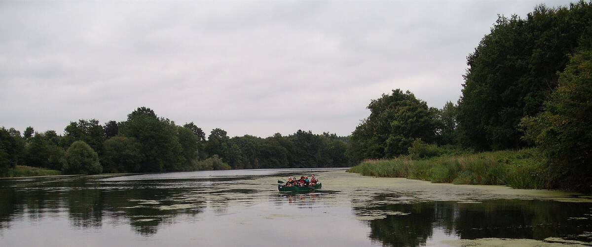 A calm river with a small boat carrying people, surrounded by dense greenery and trees.