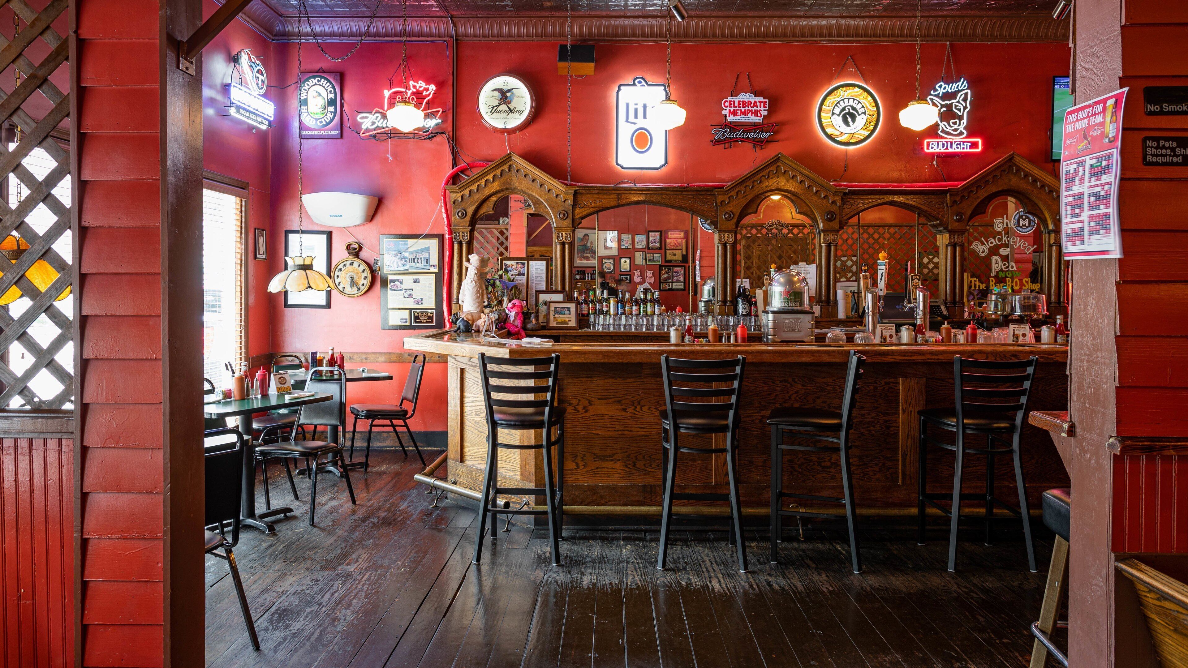 Um bar com paredes vermelhas, banquetas de madeira e diversos cartazes de cerveja e bebidas alcoólicas.