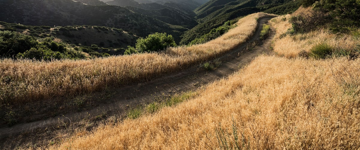 A winding mountain path through a valley with lush greenery and distant hills.