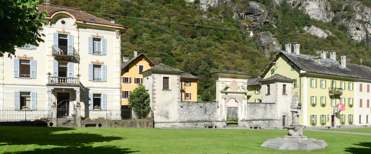 A historic town with colorful buildings, a fountain, and a mountainous backdrop.