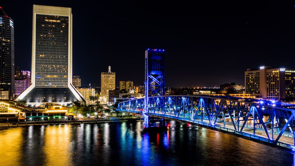 A cityscape at night with a prominent bridge illuminated in blue, a large building with a unique triangular structure, and a river reflecting the lights.