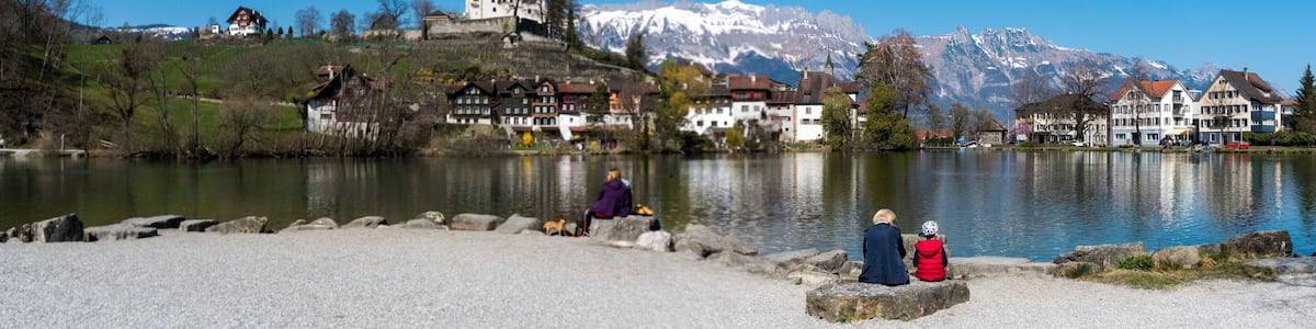 Ein See mit Häusern und einer Burg auf einem Hügel, umgeben von Bergen.