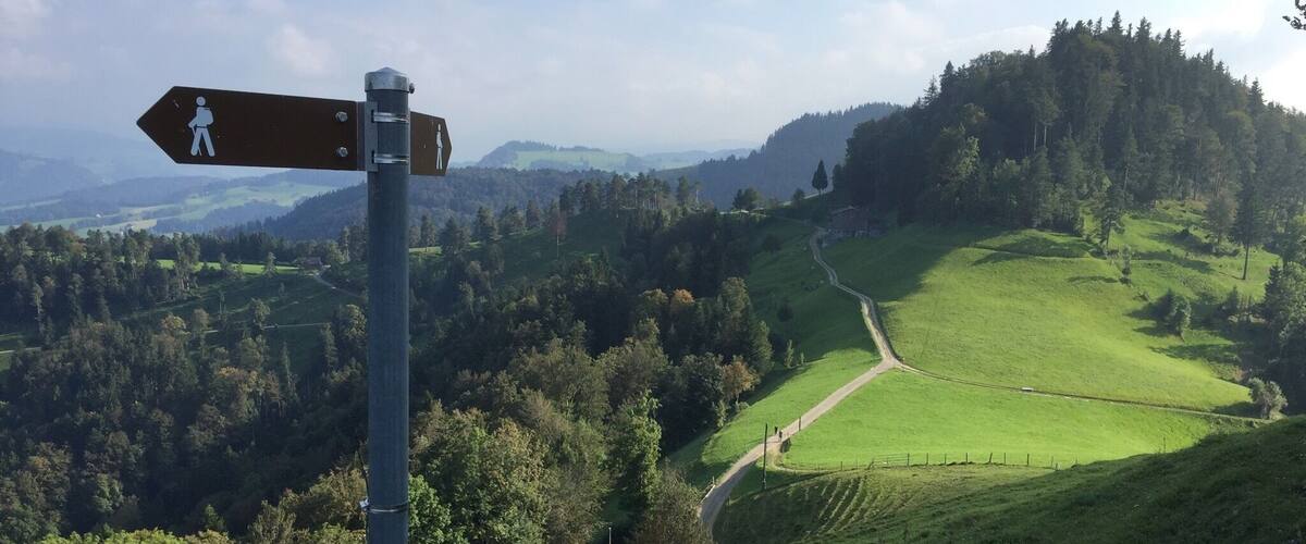 A hiking trail sign in a lush green valley with rolling hills and distant mountains.