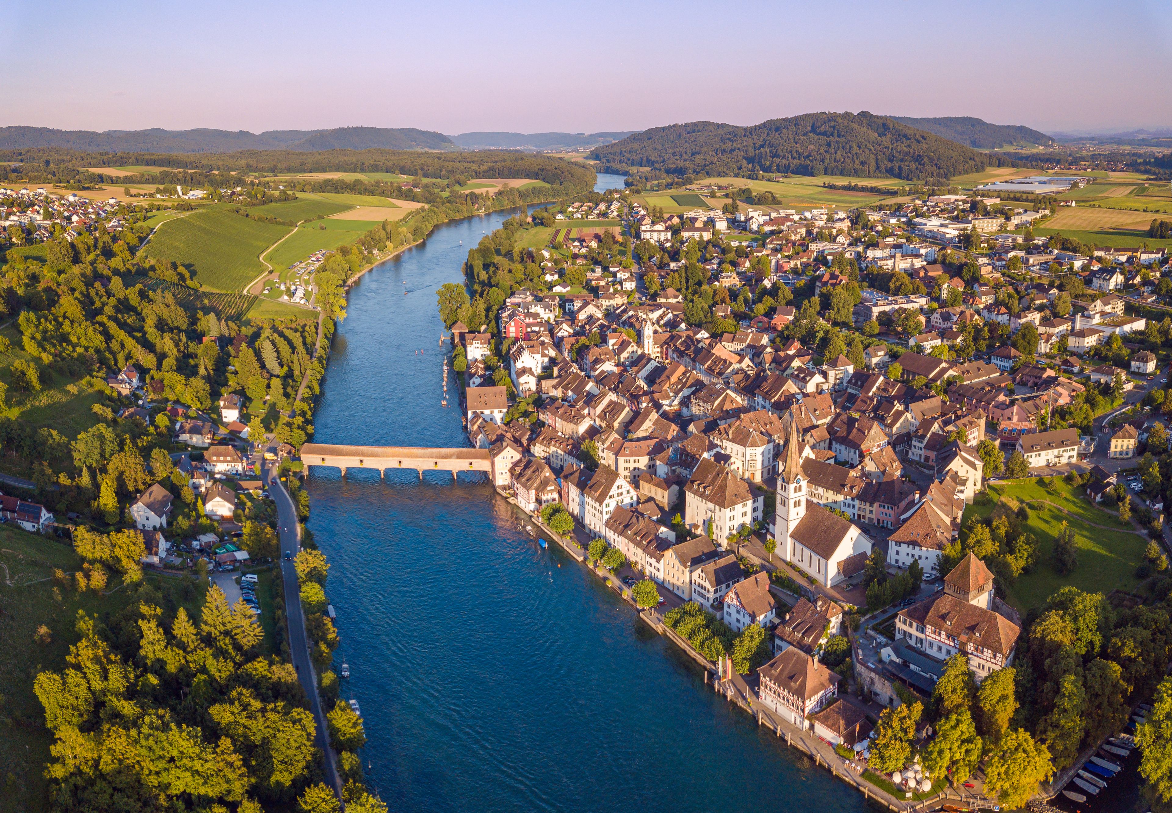 Une rivière traverse une ville où se trouve un pont, le tout étant entouré de verdure et d’immeubles d’habitation.
