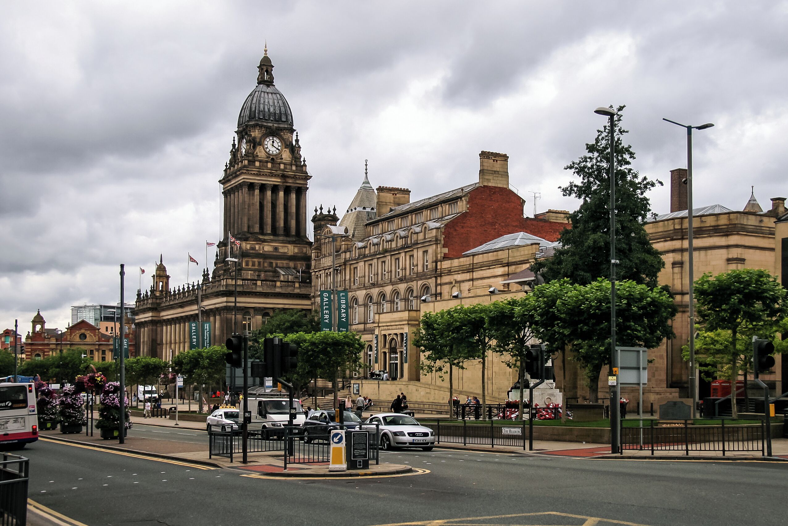 A historic building with a clock tower, a library, and a busy street with cars and a bus.