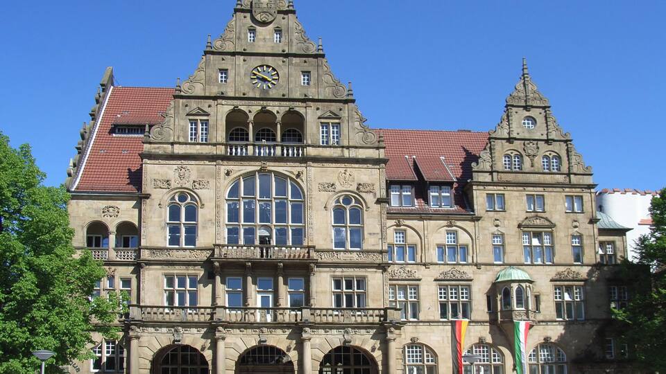 A historic building with a prominent clock tower, large arched windows, and a red-tiled roof.