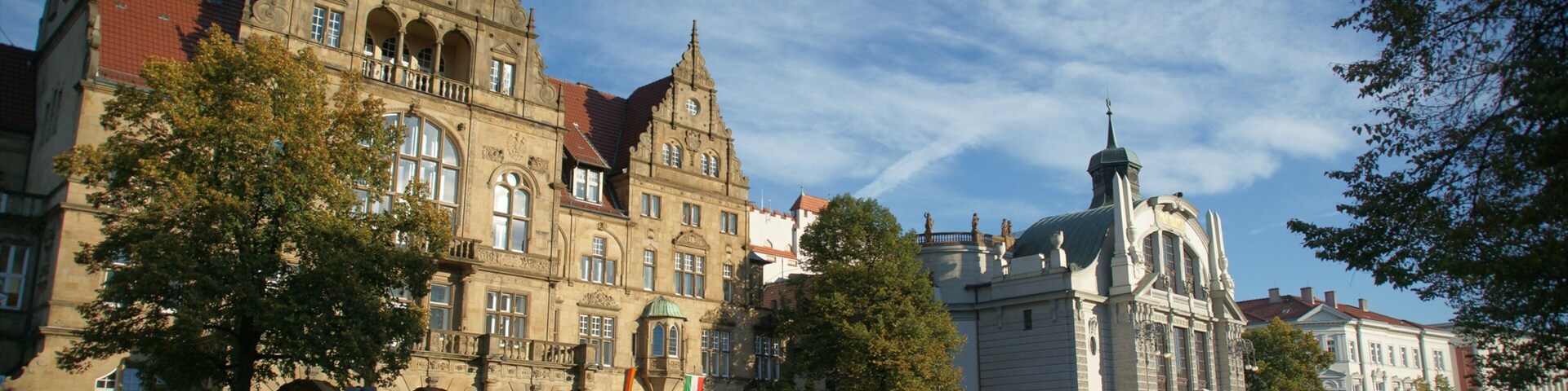 Uma praça histórica com edifícios ricamente decorados, uma fonte e pessoas caminhando.