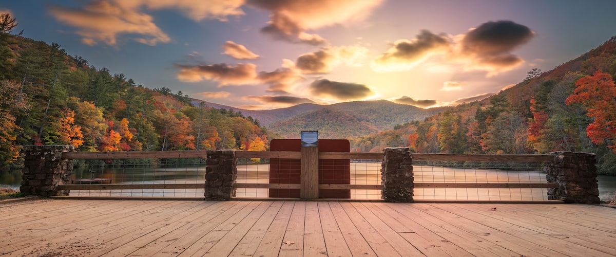 A wooden deck overlooking a lake with autumn foliage and a sunset.