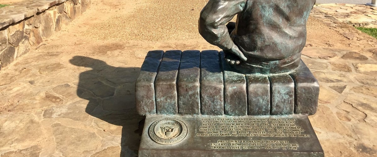 A bronze statue of a seated man with a plaque, overlooking a scenic landscape with trees and a road.