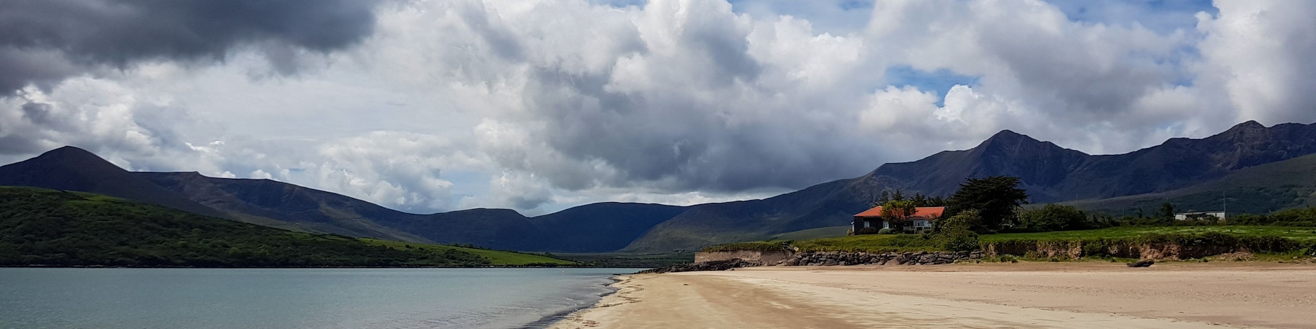 Una playa con huellas que conducen al agua, montañas al fondo y una casa con techo rojo.