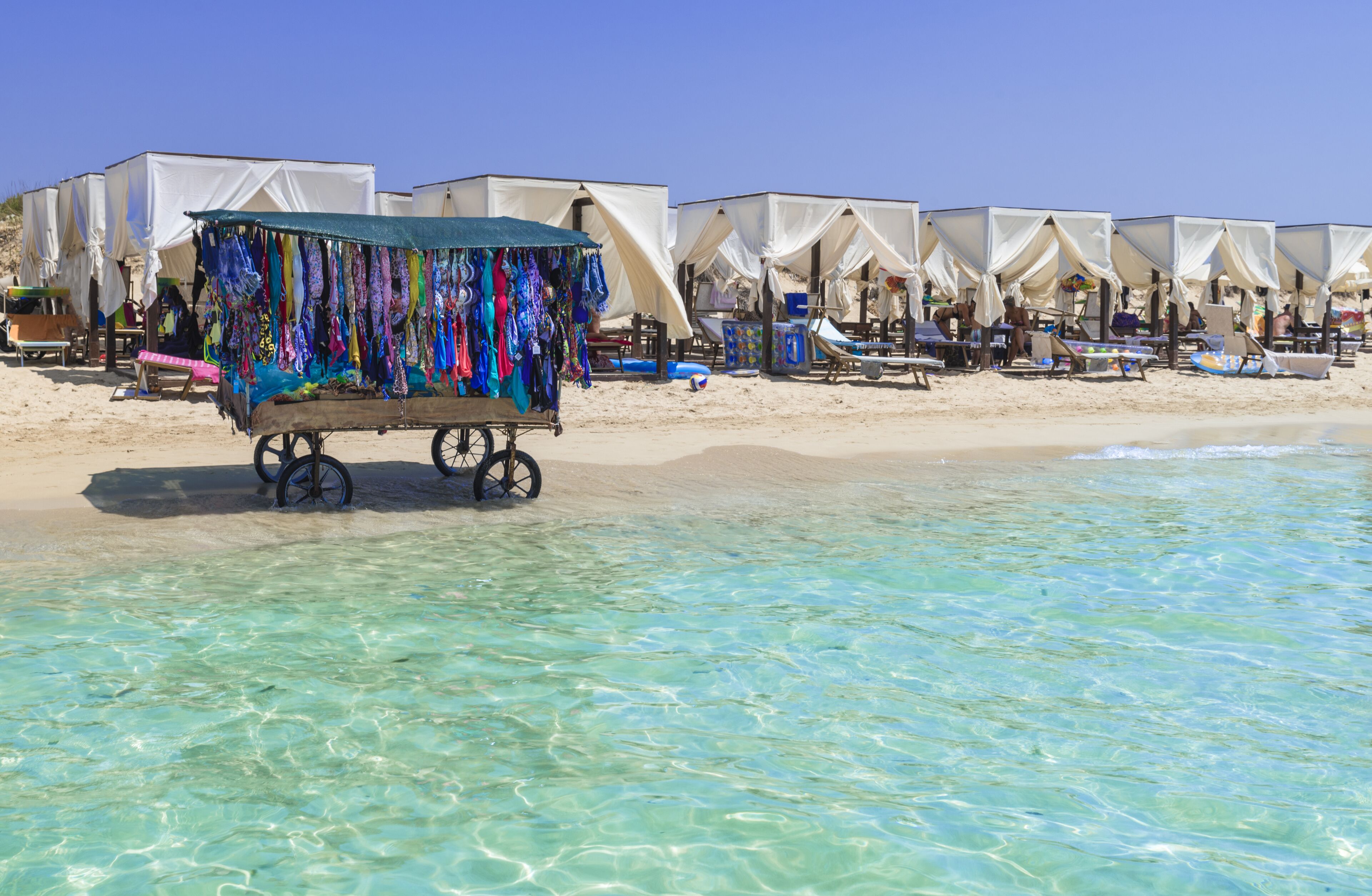 A beach with clear turquoise water, white tents, and a cart selling colorful clothing.