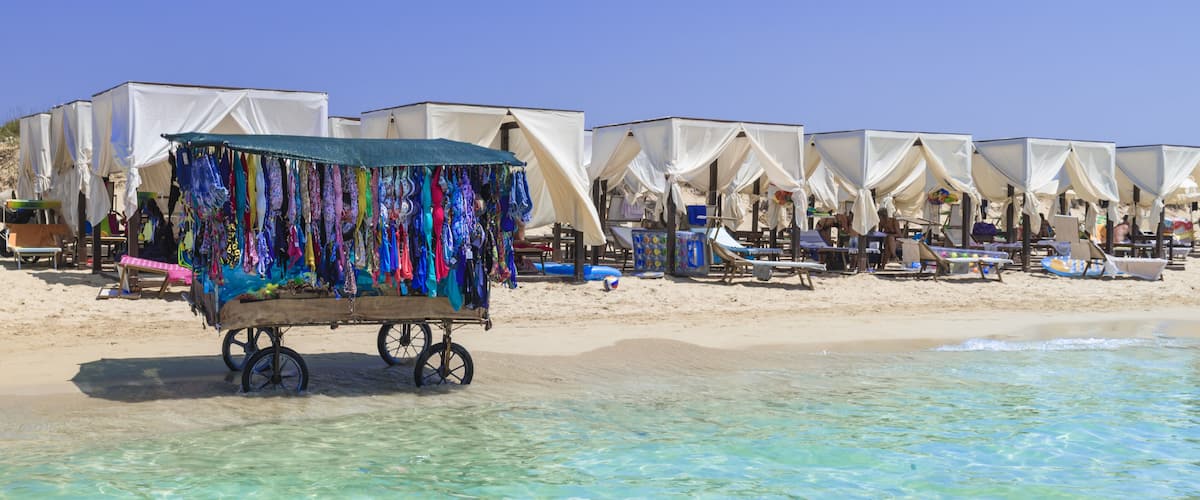 A beach with clear turquoise water, white tents, and a cart selling colorful clothing.