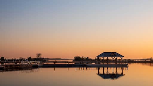 A tranquil lakeside scene at sunset with a wooden pavilion on stilts and a calm water surface reflecting the sky.