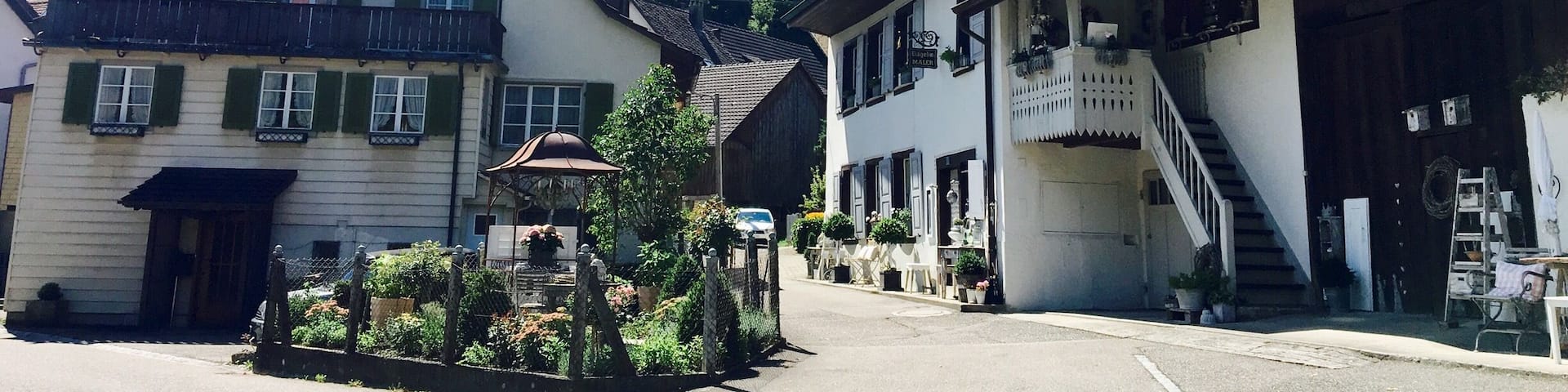 A quaint European village street with traditional buildings, a small fountain, and a clear blue sky.