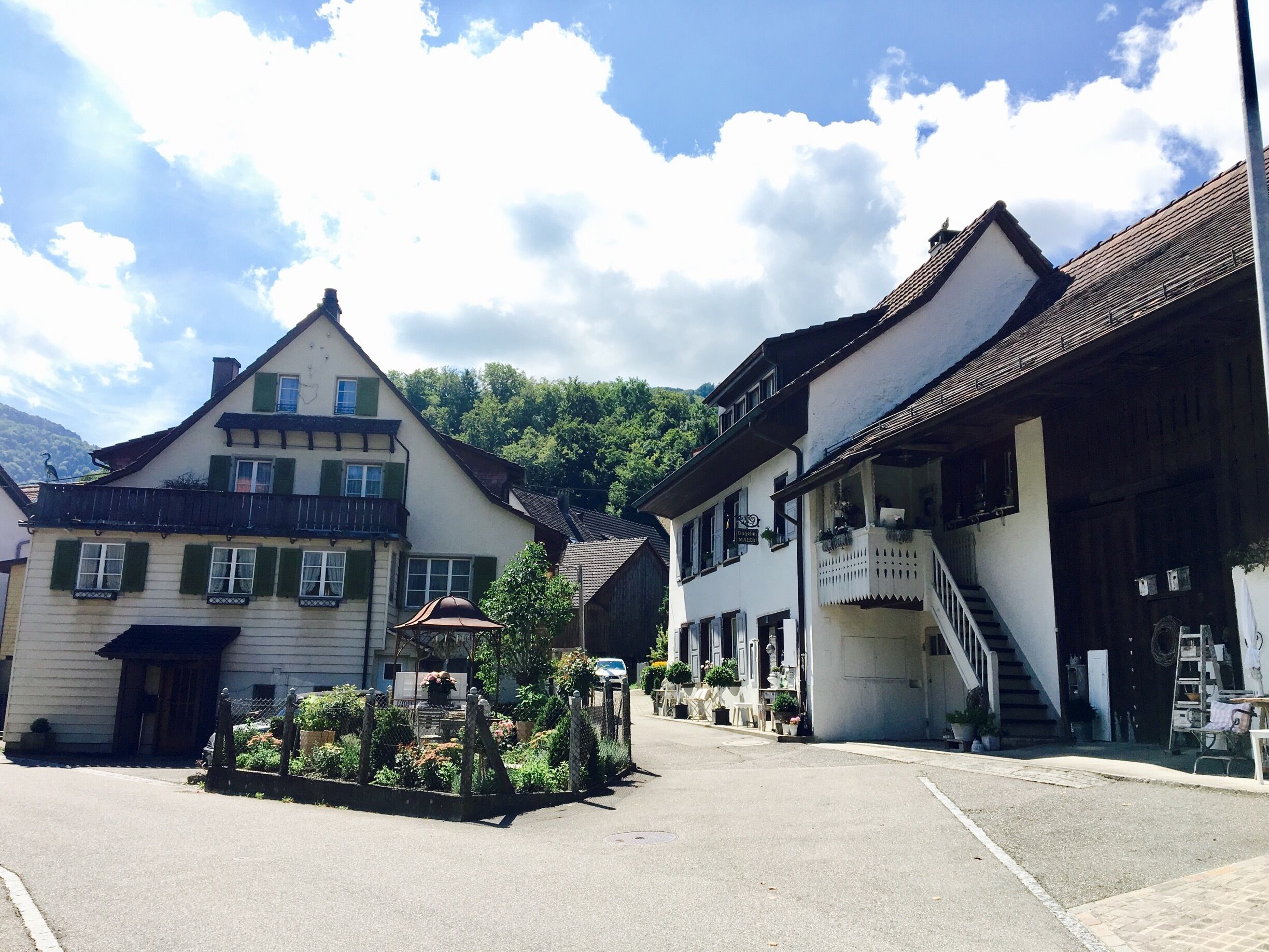 A quaint European village street with traditional buildings, a small fountain, and a clear blue sky.