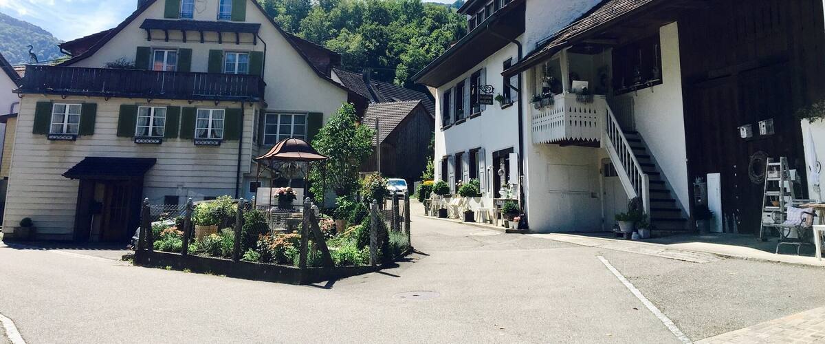 A quaint European village street with traditional buildings, a small fountain, and a clear blue sky.