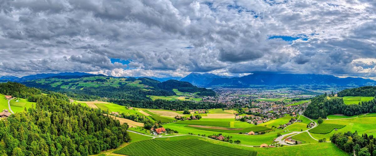 A panoramic view of a lush valley with green fields, a small town, and distant mountains under a cloudy sky.
