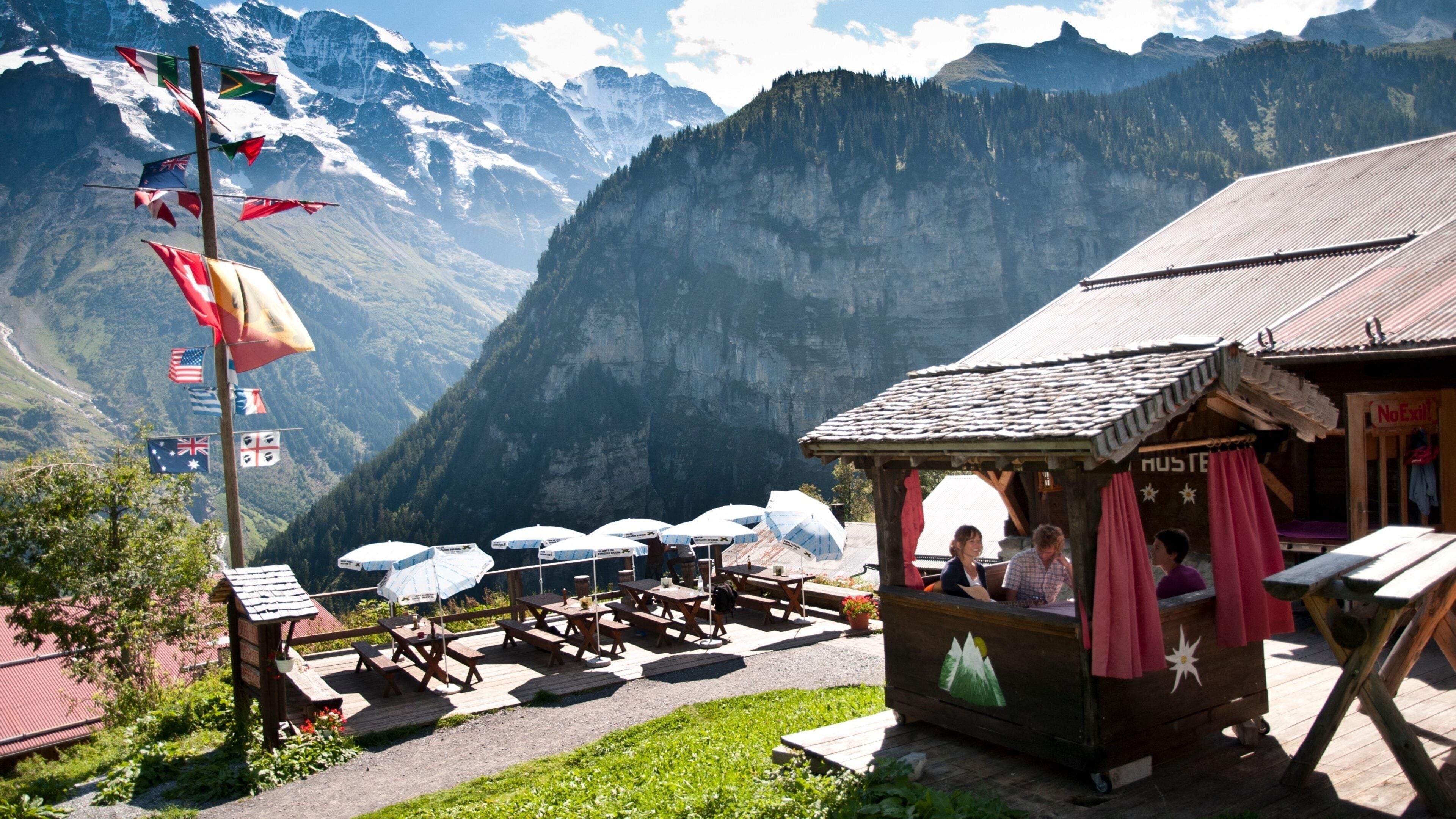 A mountain lodge with picnic tables and umbrellas, surrounded by flags and a view of snow-capped mountains.