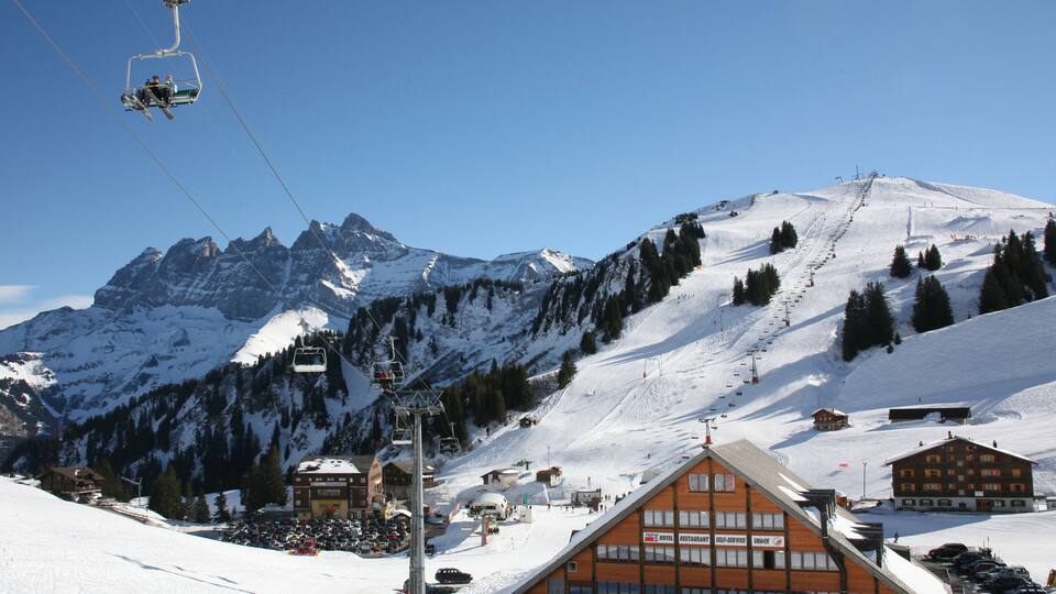 A ski lift, snow-covered mountains, a ski resort building, and a clear blue sky.