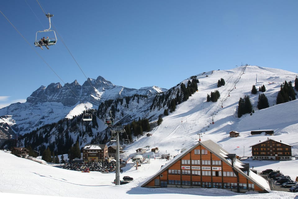 A ski lift, snow-covered mountains, a ski resort building, and a clear blue sky.