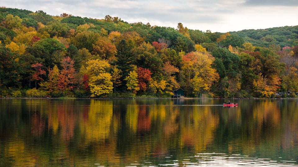 A calm lake with autumn foliage reflecting on the water, surrounded by a forested hillside.