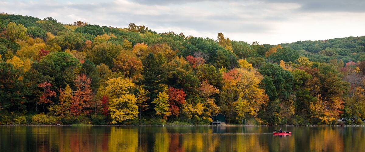 A calm lake with autumn foliage reflecting on the water, surrounded by a forested hillside.