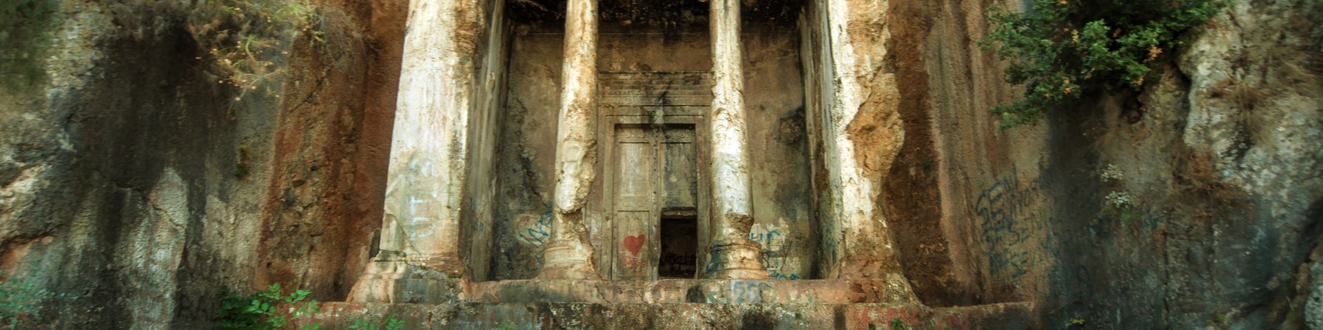 Um templo antigo com colunas e uma porta central, cercado por terreno rochoso e vegetação.