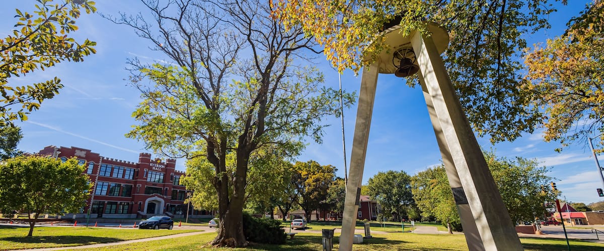A park with a large stone arch, trees, and a red brick building in the background.