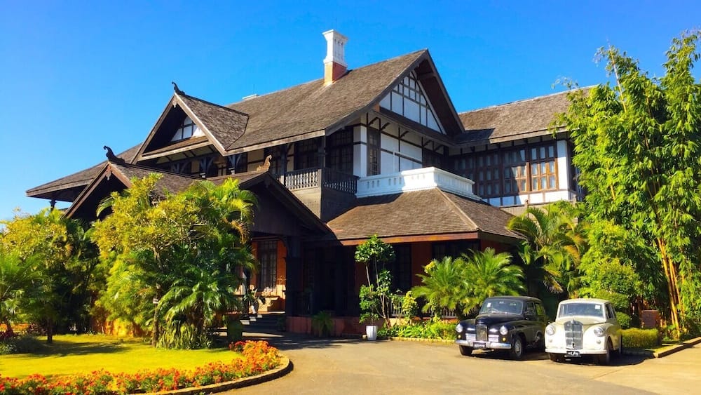 A large, traditional house with a steeply pitched roof, surrounded by lush greenery and palm trees.