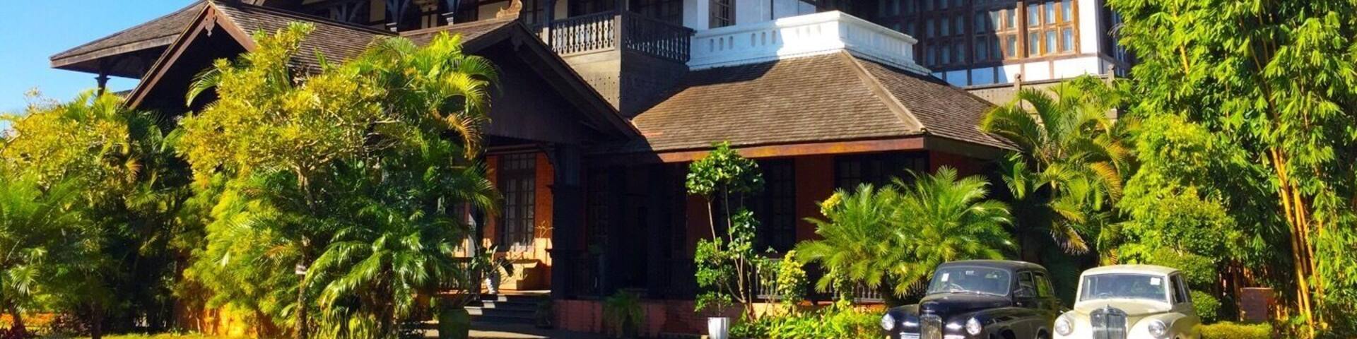 A large, traditional house with a steeply pitched roof, surrounded by lush greenery and palm trees.