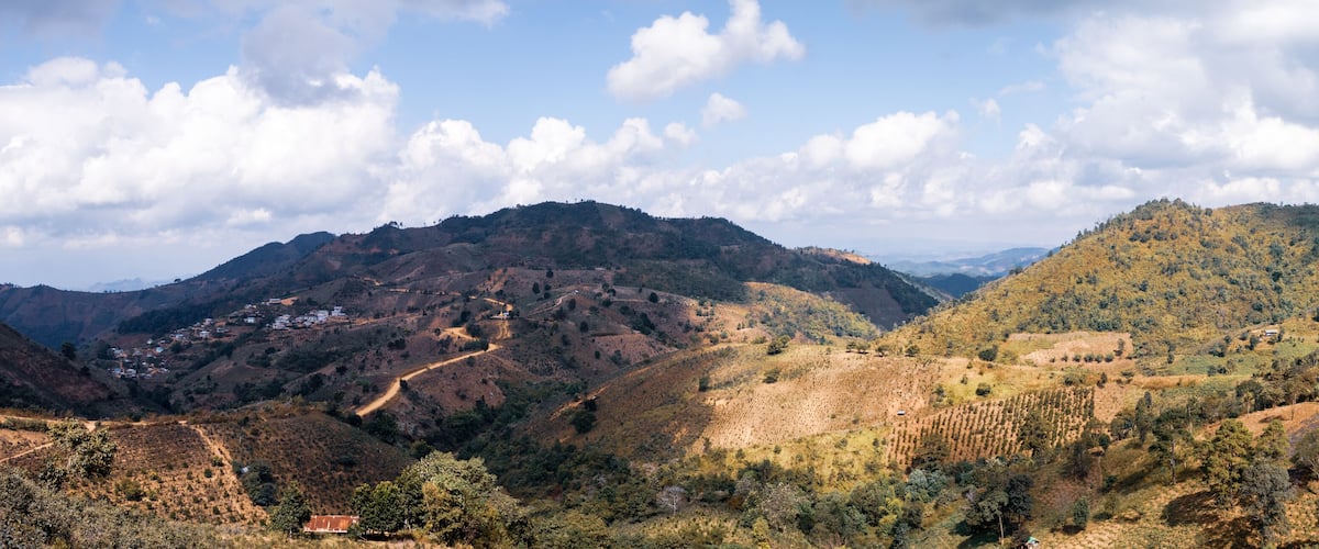 A mountainous landscape with a winding road, scattered houses, and dense forested areas.