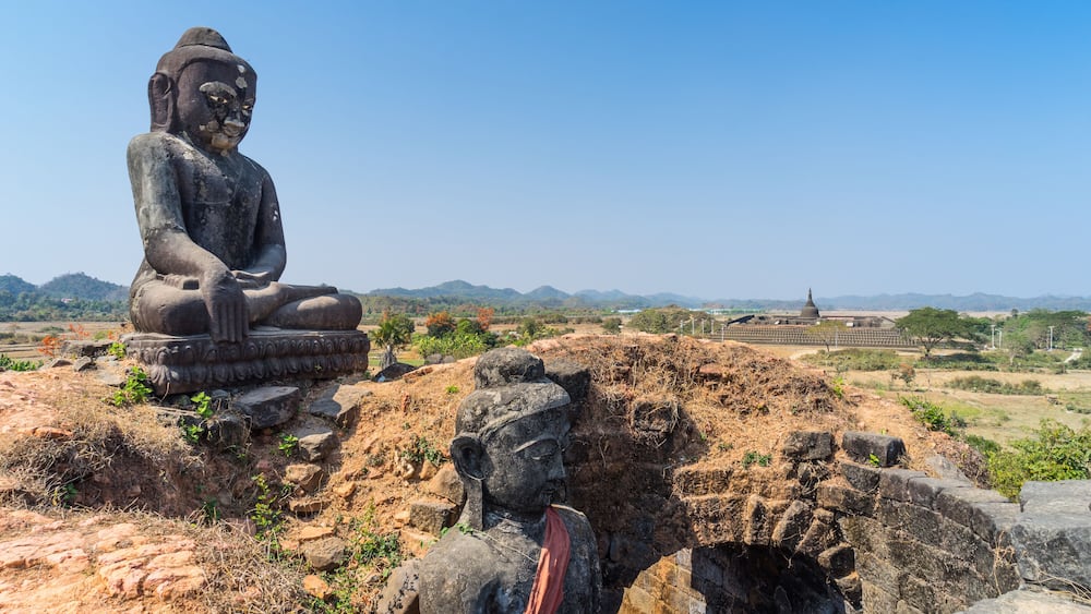 A large seated Buddha statue on a pedestal, surrounded by dry grass and rocks, with a clear blue sky and distant hills in the background.