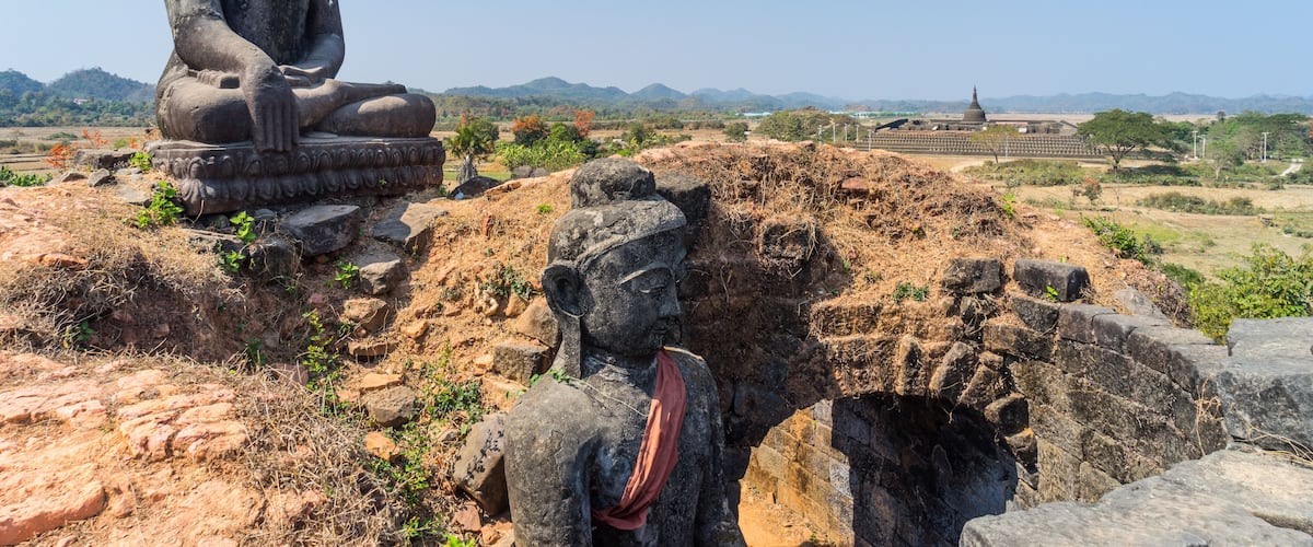 A large seated Buddha statue on a pedestal, surrounded by dry grass and rocks, with a clear blue sky and distant hills in the background.