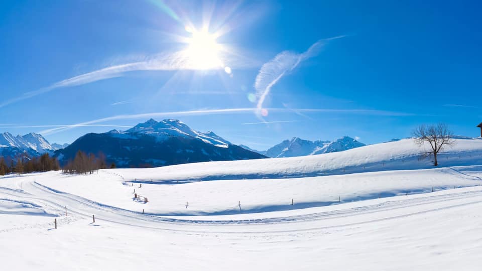 A snow-covered ski slope with a wooden cabin, a ski lift, and a mountain range under a clear blue sky.