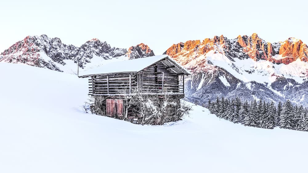 Une cabane en bois, située sur le versant enneigé d’une montagne, avec des arbres couverts de neige et une chaîne de montagnes en arrière-plan.