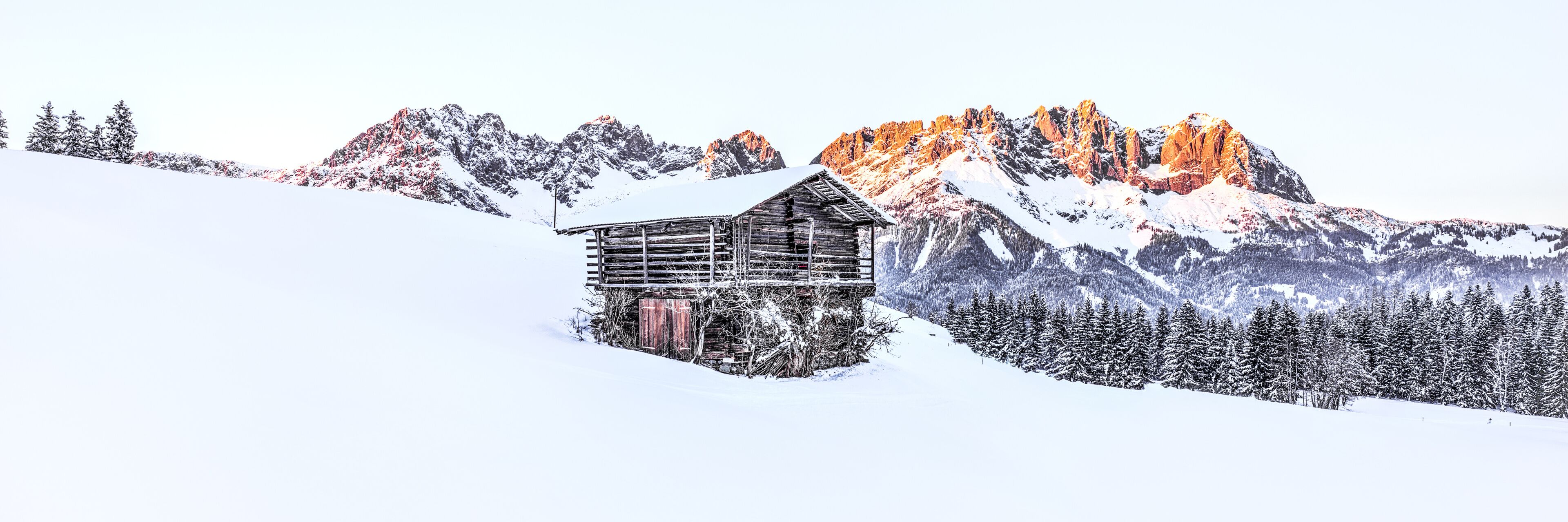 A wooden cabin on a snowy mountain slope with snow-covered trees and a range of mountains in the background.