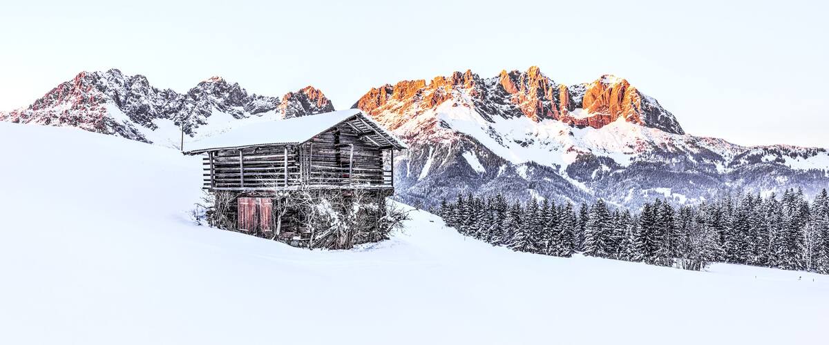 A wooden cabin on a snowy mountain slope with snow-covered trees and a range of mountains in the background.