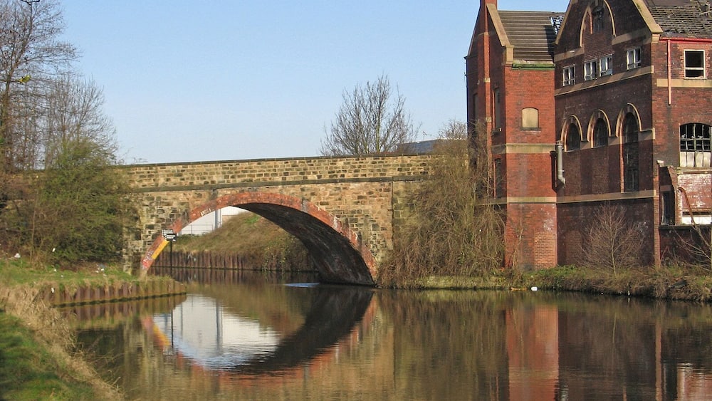 A stone bridge over a calm river, reflecting the surrounding trees and a brick building with arched windows.