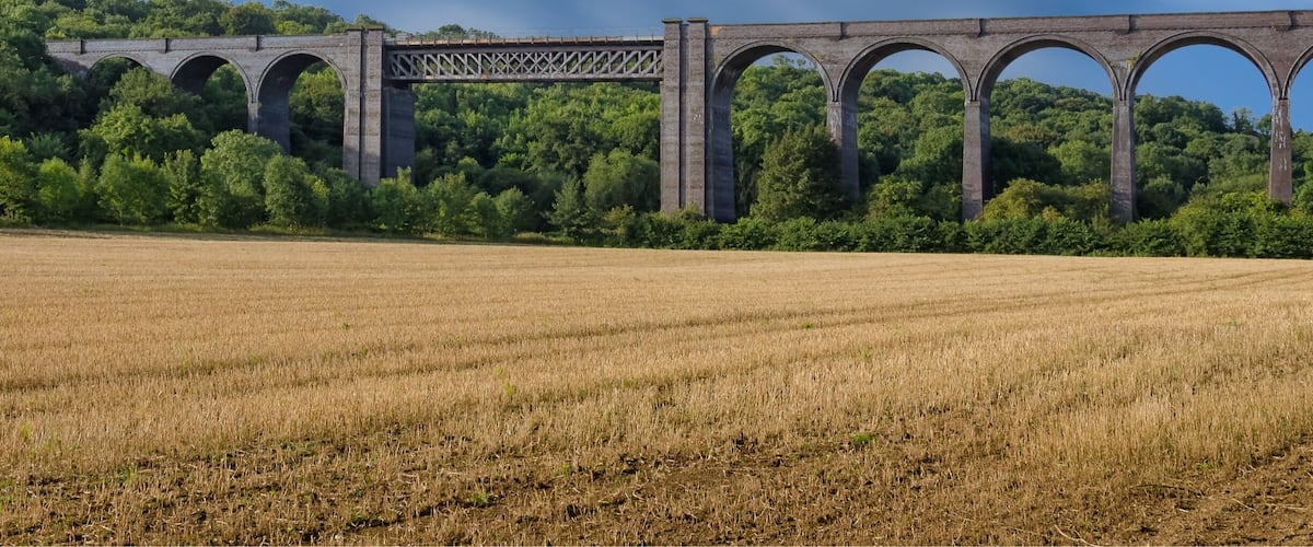 A stone viaduct over a field with a forested hill in the background.