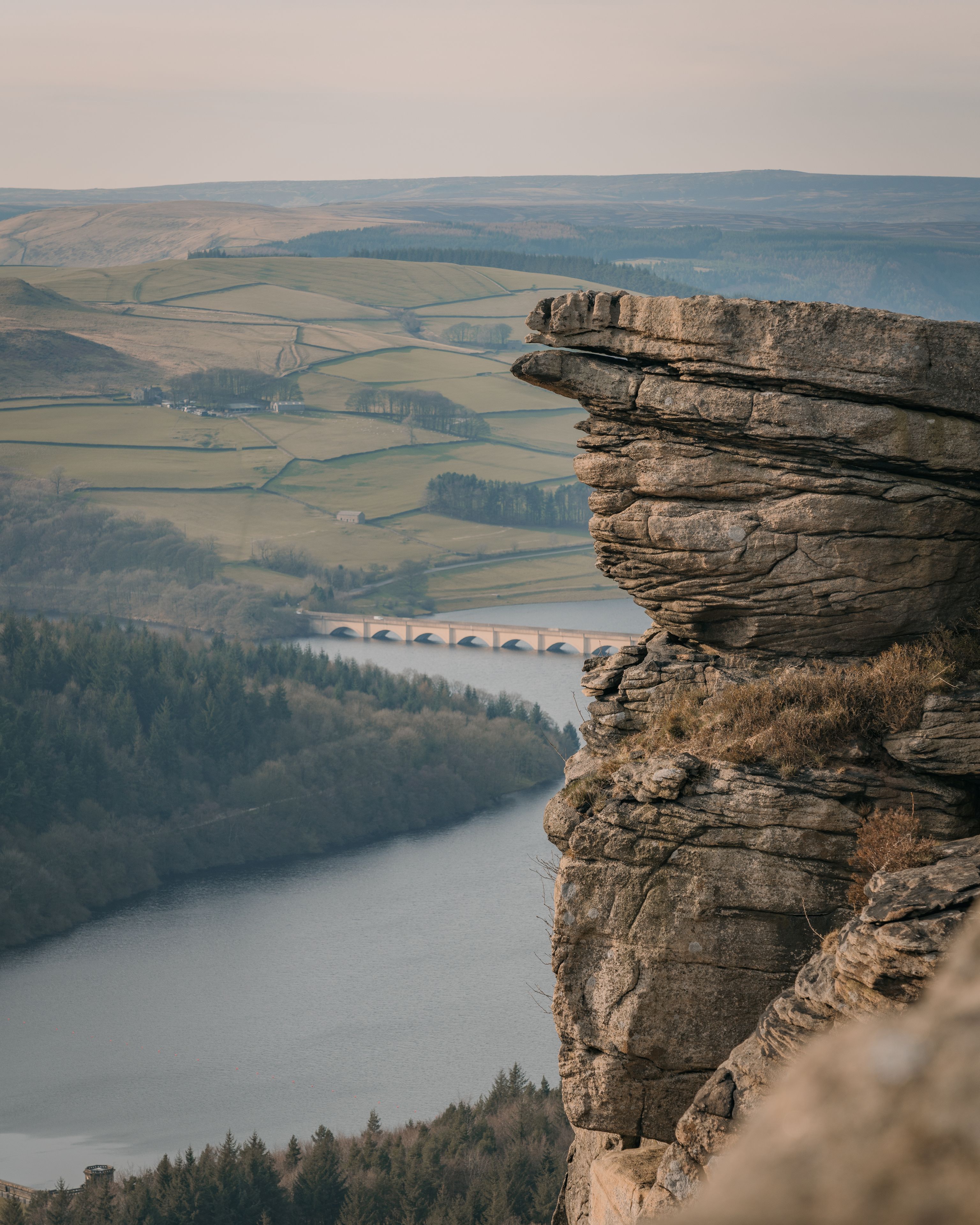 A rocky cliff overlooking a river with a bridge in the distance.