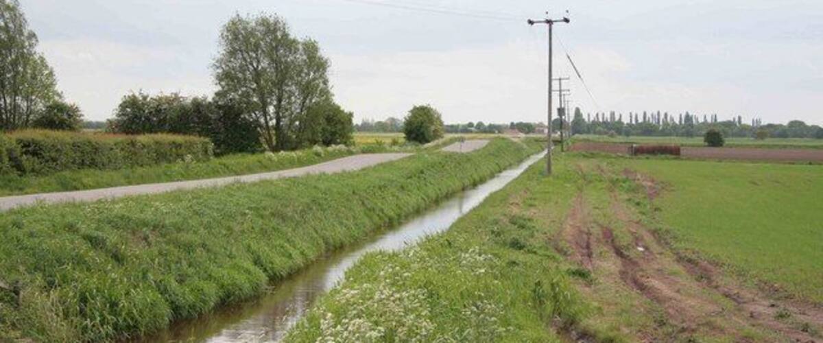 A narrow water channel flanked by grassy fields and a dirt path.